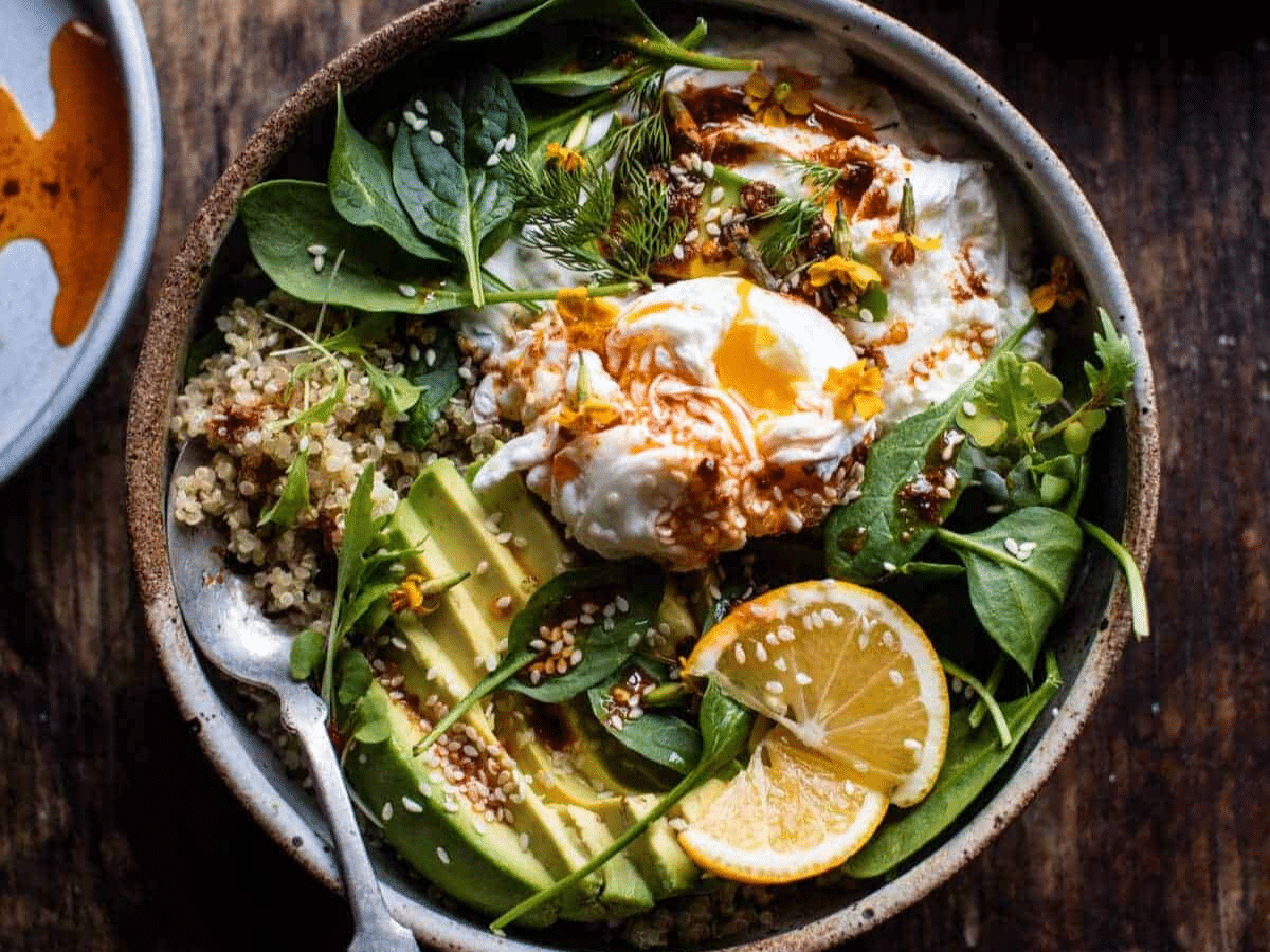 A quinoa breakfast bowl with poached eggs, avocado slices, greens, and lemon slices.