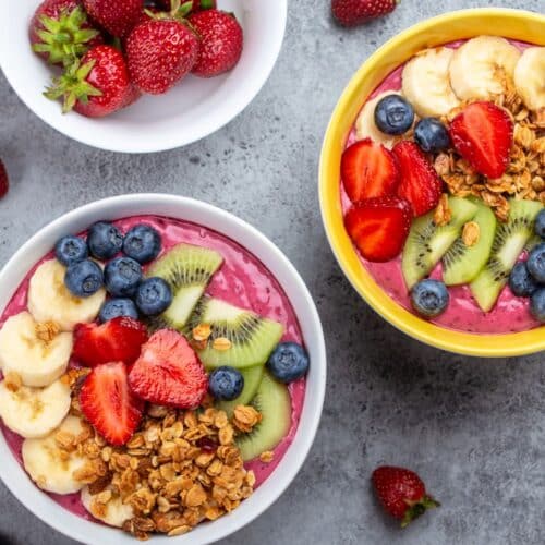 Two pink smoothie bowls topped with bananas, strawberries, kiwi, bluberries, next to a bowl of whole strawberries.