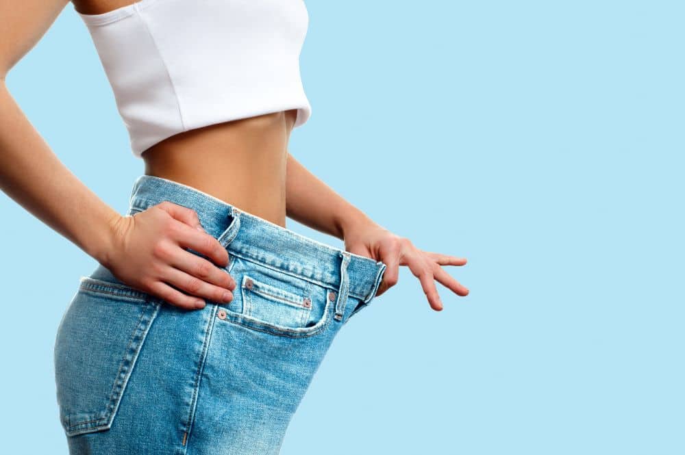 A woman in oversized jeans showing her weight loss against a blue background.