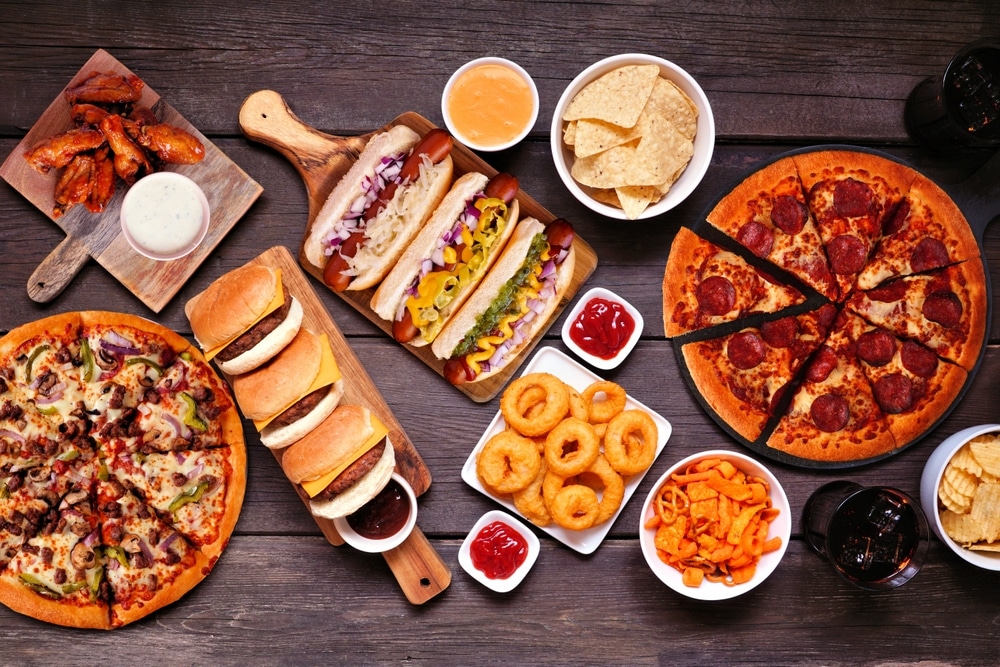 A table of pizza, burgers, hot dogs, onion rings, and other junk food.