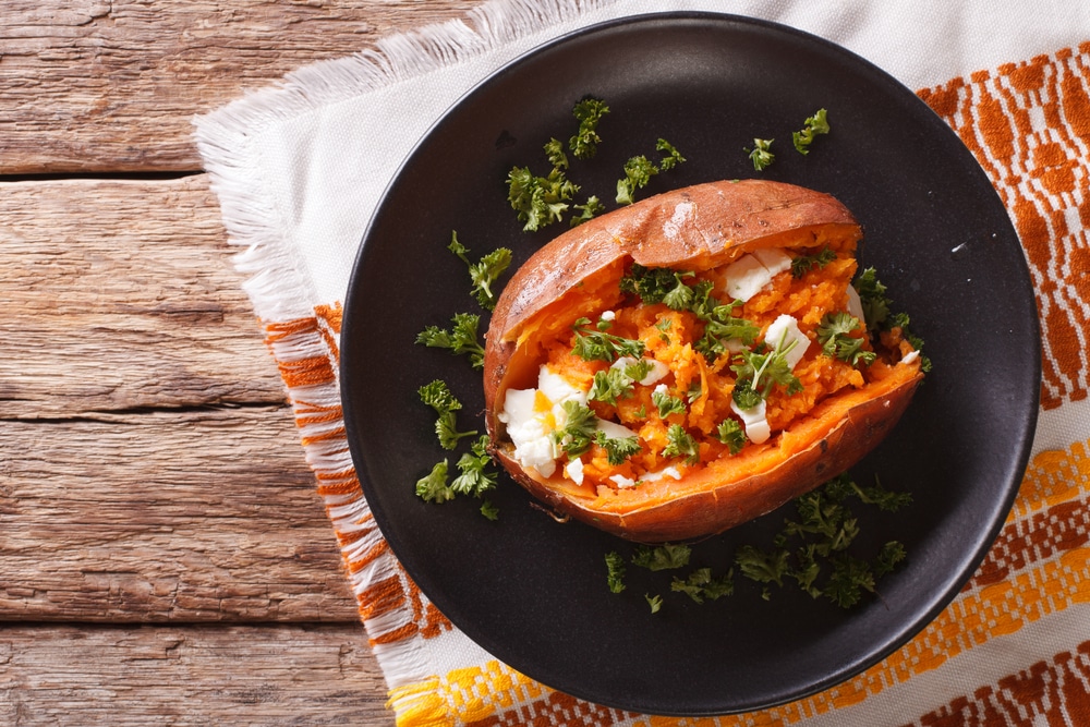Whole baked sweet potato on a black plate with cheese, spices, and parsley.
