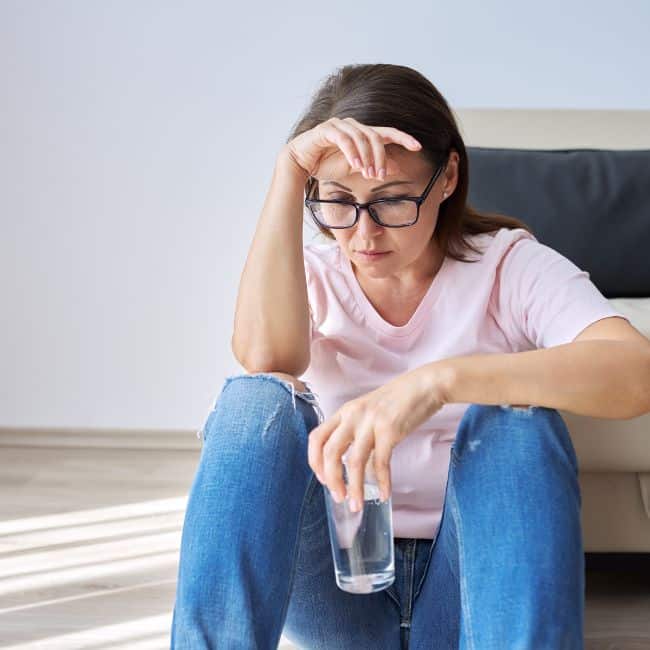 woman sitting on floor looking fatigued holding glass of water with hand on forehead