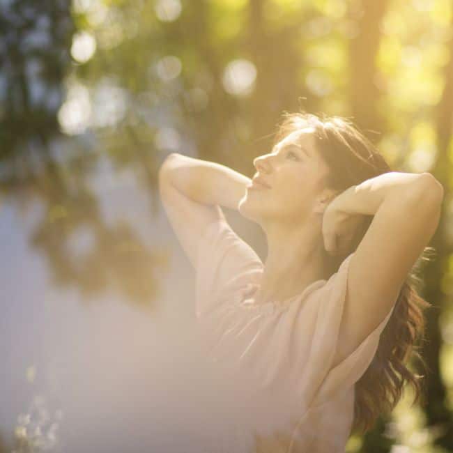 woman standing in sunlight with arms behind her head smiling at sunshine