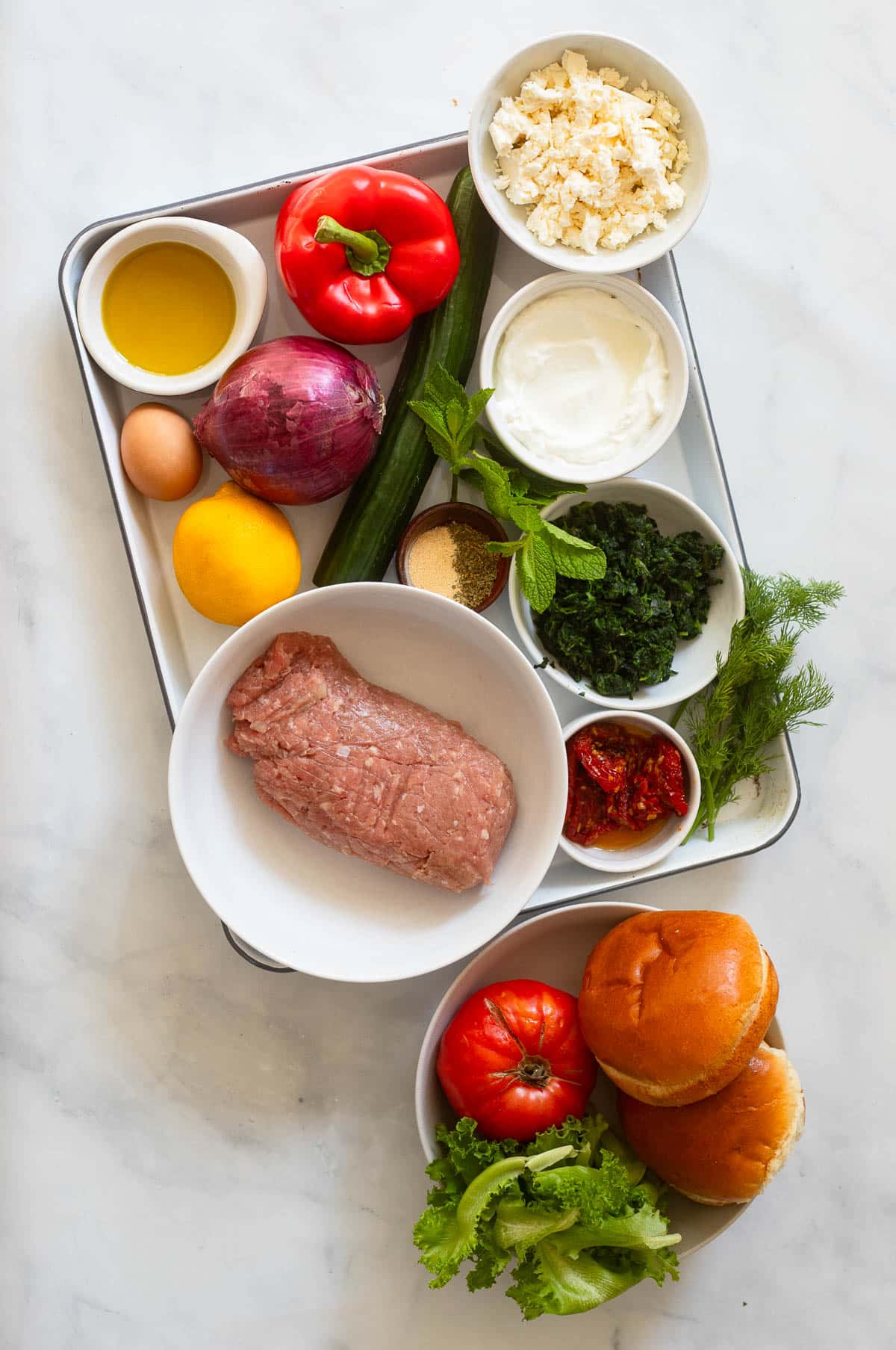 top view of raw greek turkey burger ingredients in bowls and on tray on white counter backdrop