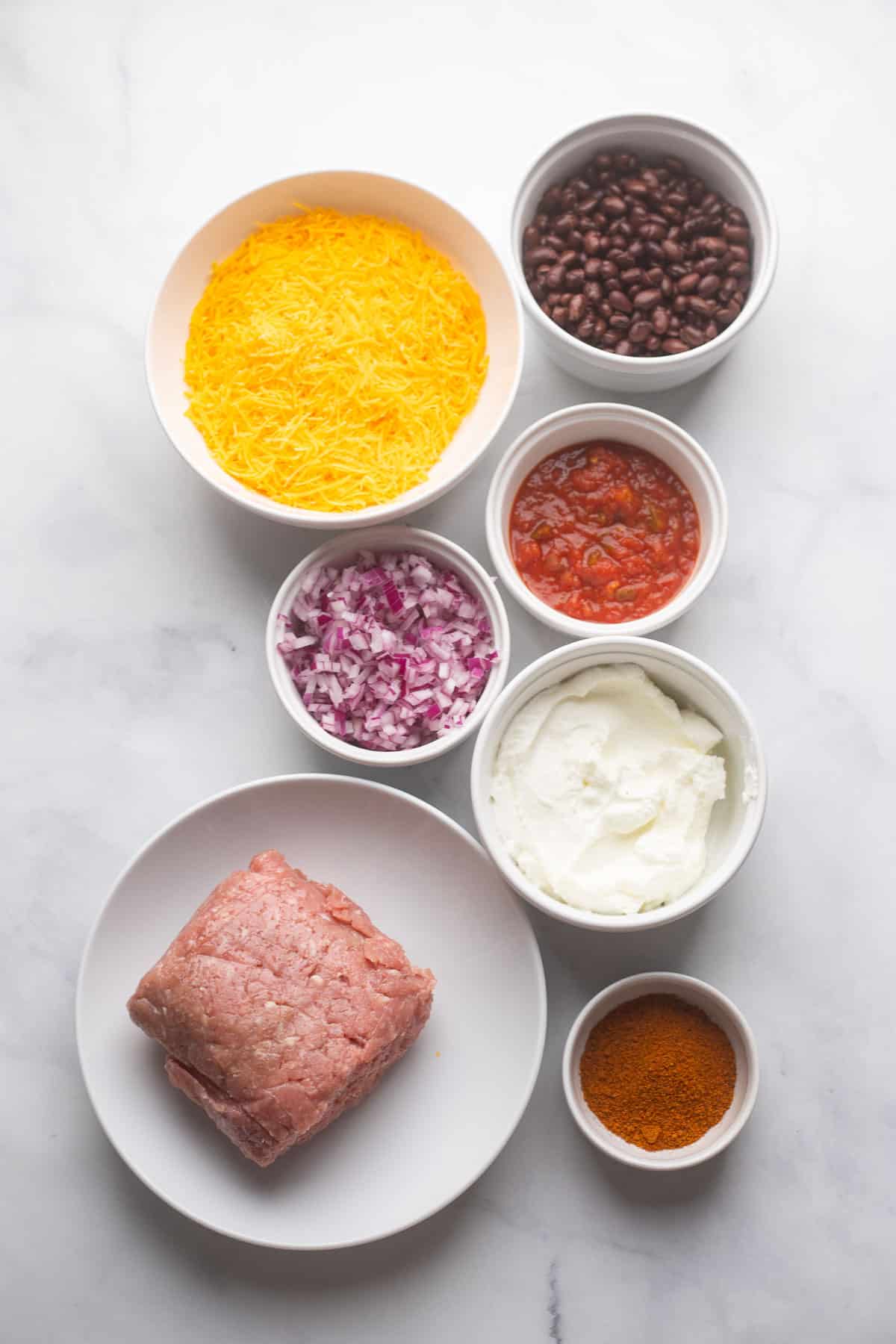 healthy chili cheese dip ingredients separated in white bowls on white counter backdrop