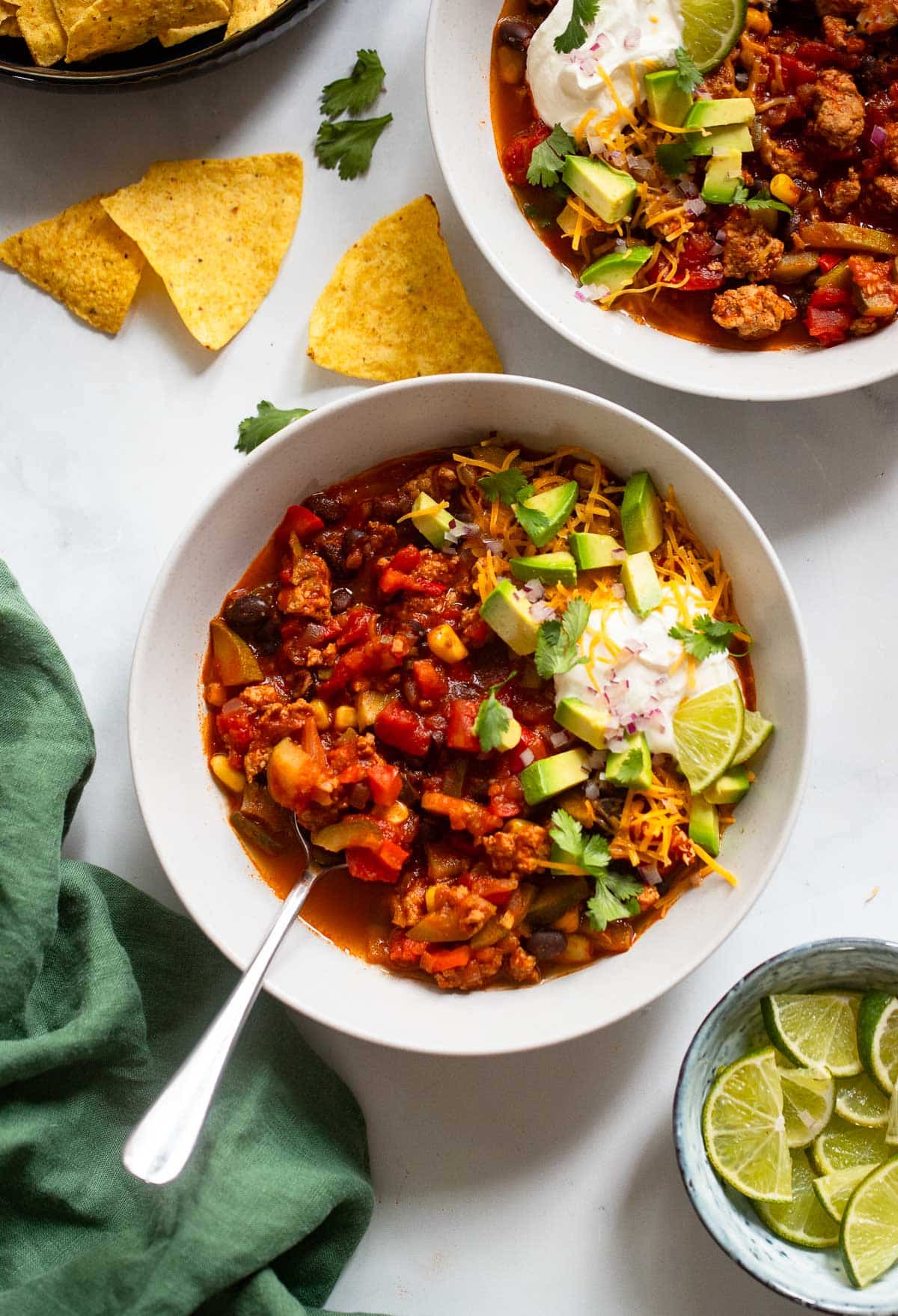 two individual servings of healthy turkey chili with spoon on white counter with topping sides