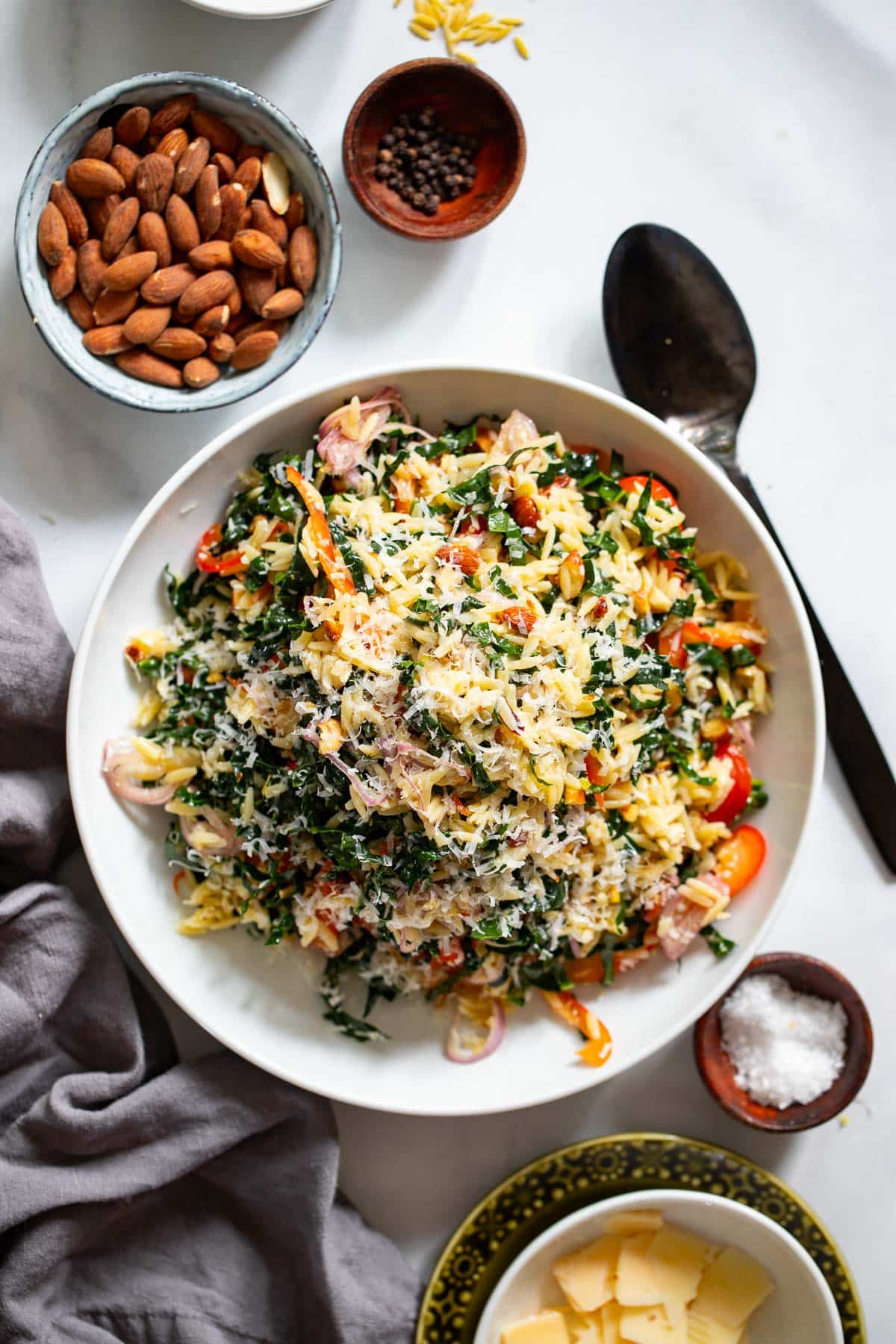 serving of orzo kale salad in white bowl on white counter with toppings laid out around
