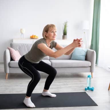 Older woman doing squats on a black yoga mat in her living room.