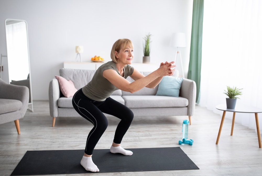 Older woman doing squats on a black yoga mat in her living room.