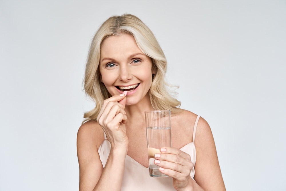 Older woman smiling while taking a dietary supplement with a glass of water.