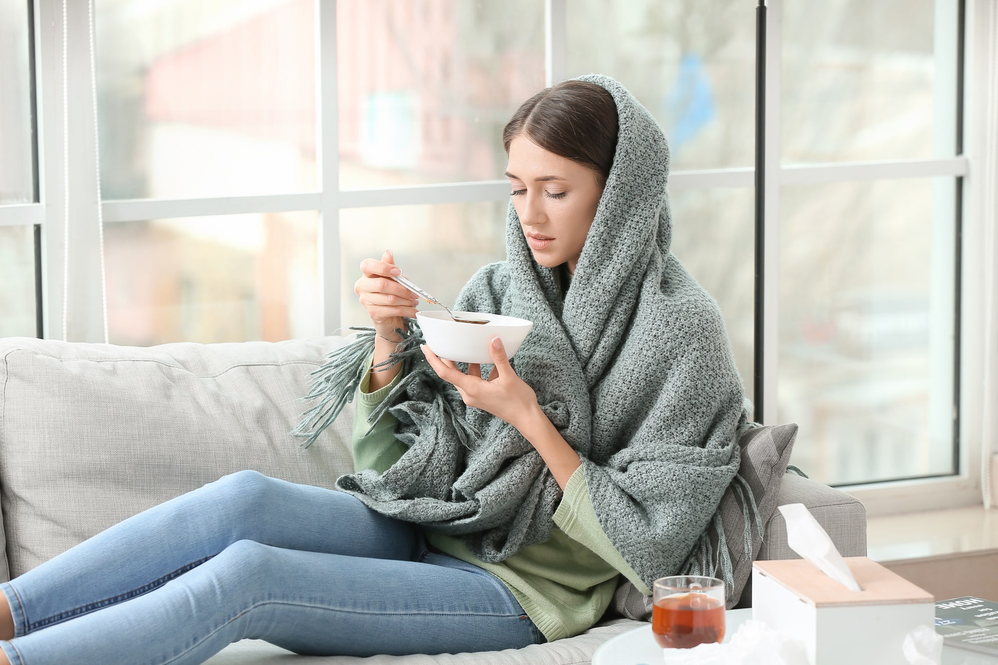 Sick young woman eating chicken soup while wrapped in a blanket on the couch.