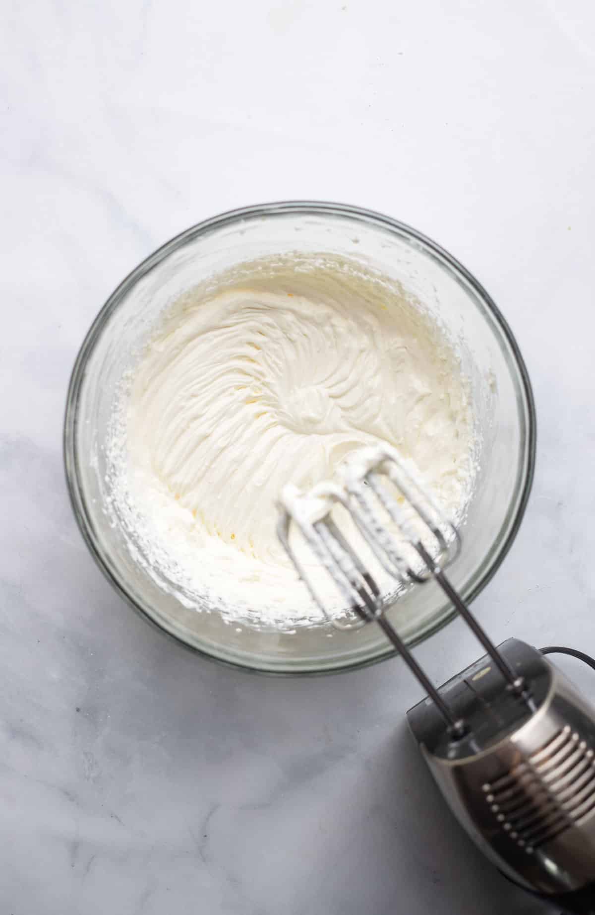 overhead view of hand mixer mixing cream cheese frosting on white countertop