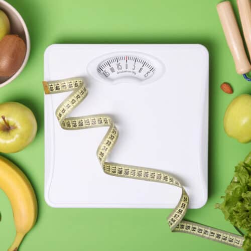 A floor scale and tape measure surrounded by healthy foods on a green background.
