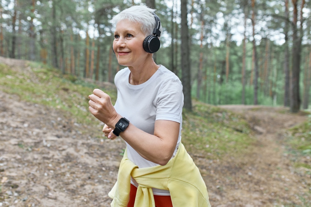 An older woman power walking in the woods with headphone on.