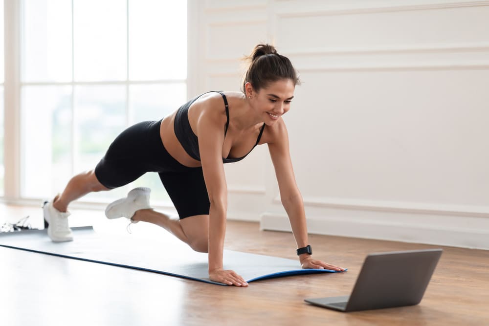 A woman doing a mountain climber exercise on a yoga mat in front of her laptop.