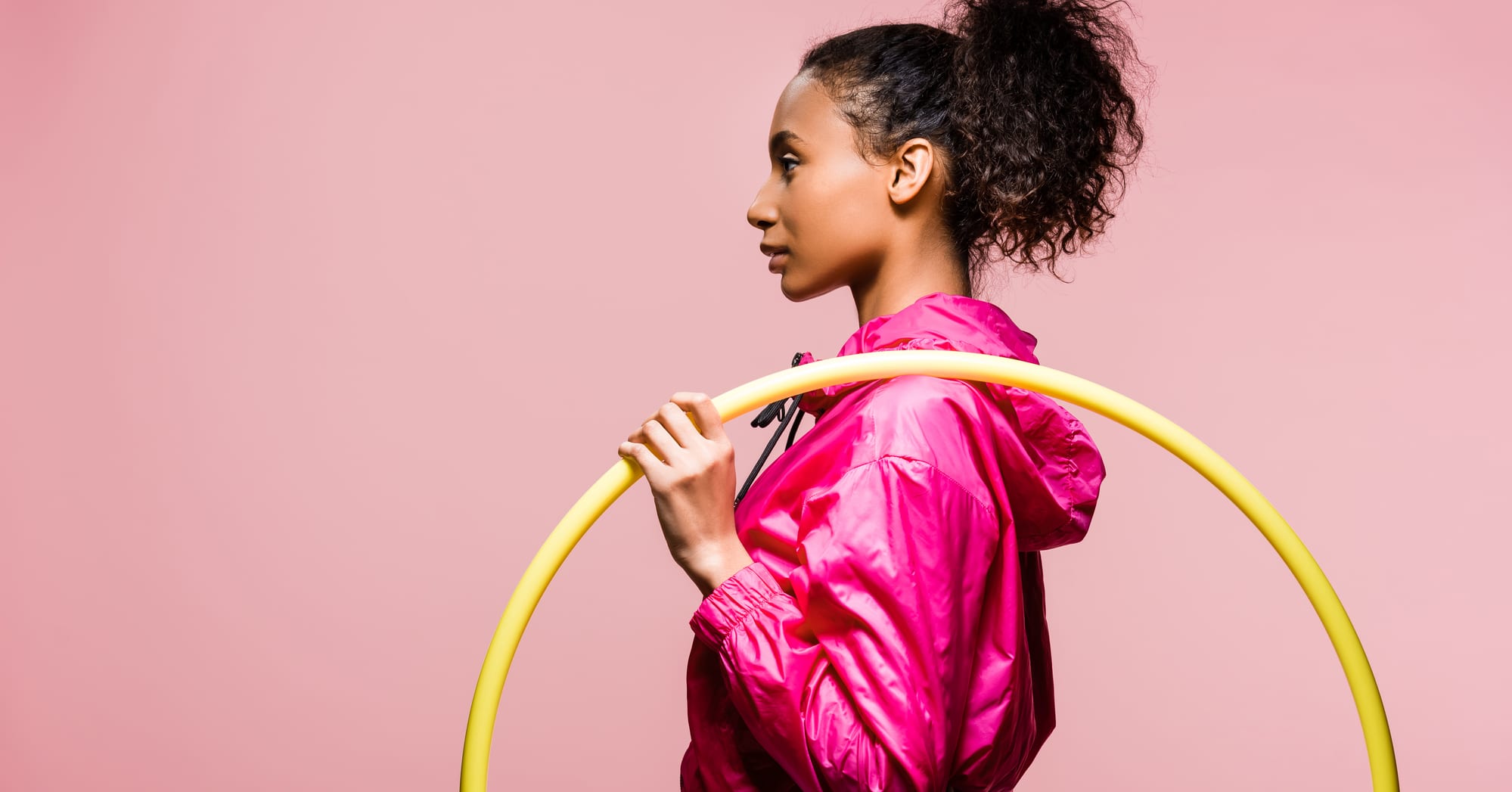 Side view of a woman in a pink jacket holding a yellow weighted hula hoop on her shoulder.