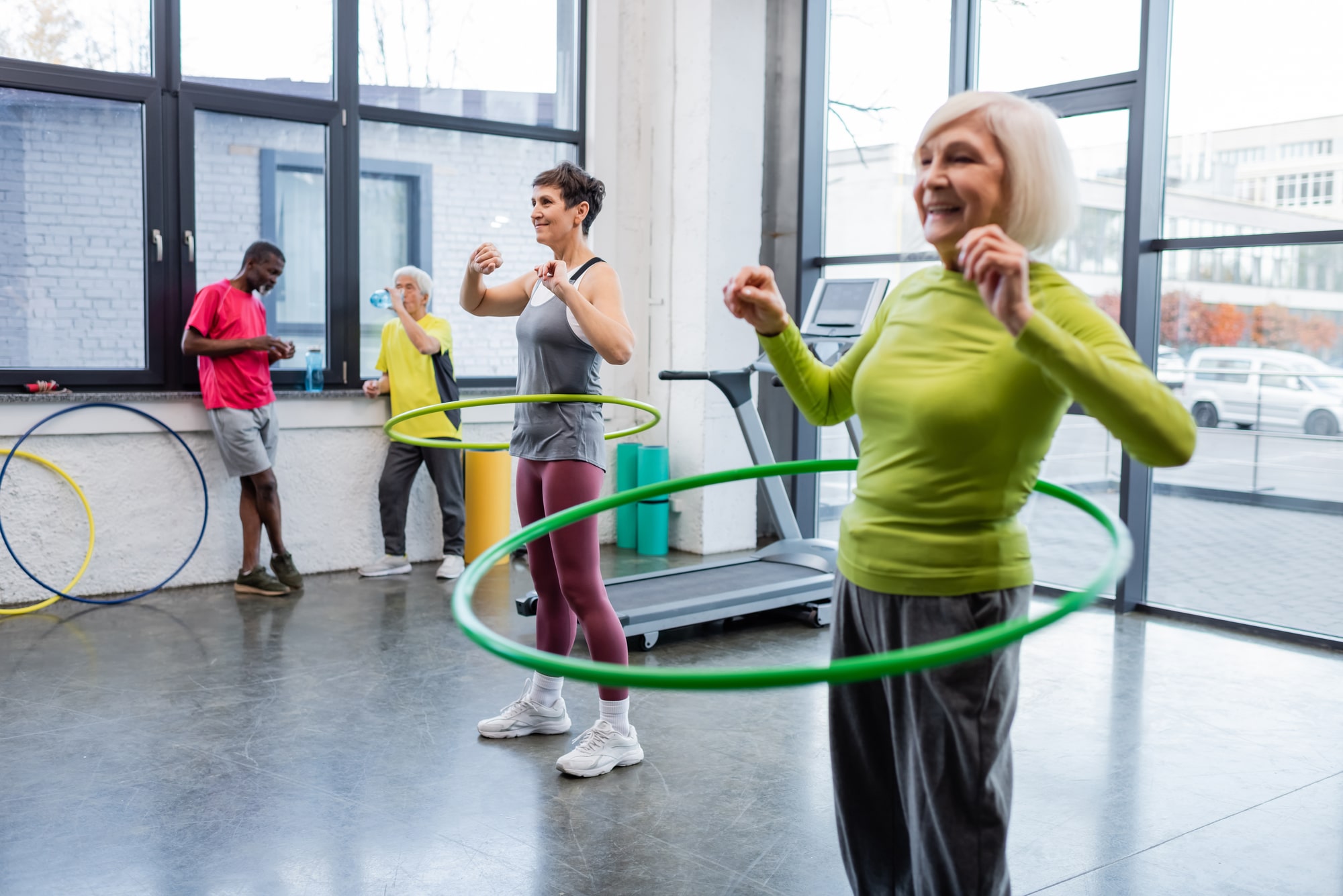 A group fitness class with a close up of an older woman exercising with a weighted hula hoop.
