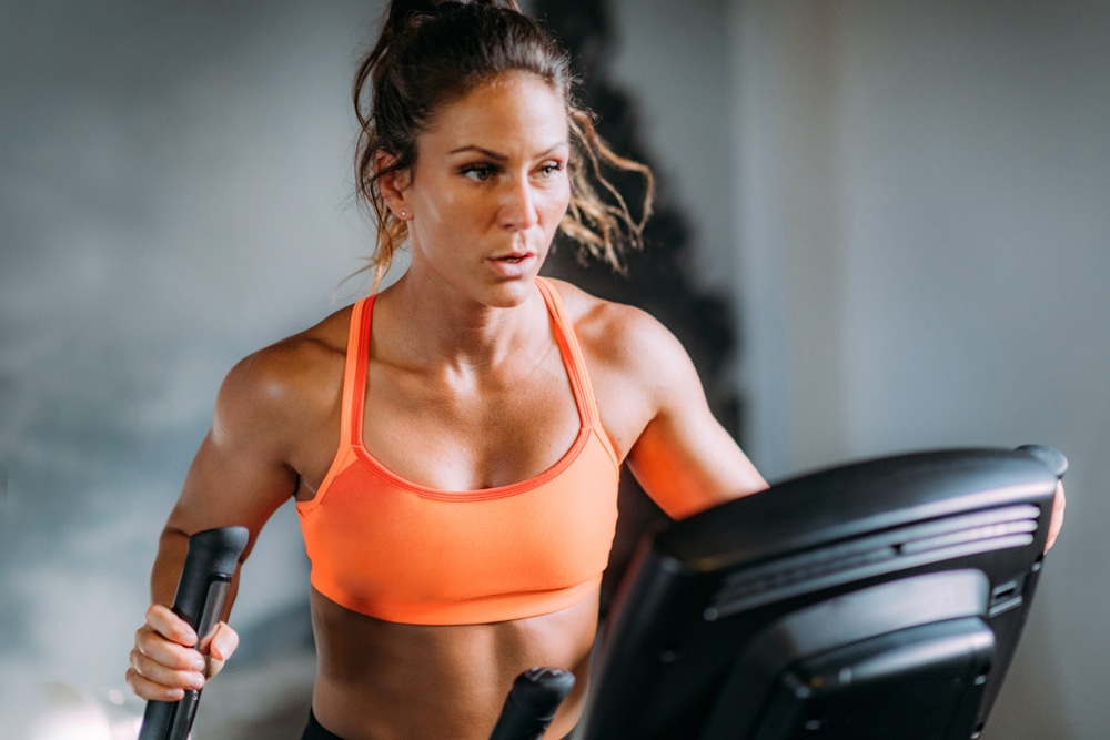 Close up of a woman in an orange workout top working out on an elliptical machine.