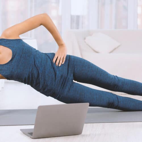 A woman doing a side plank on a grey yoga mat in front of her laptop.