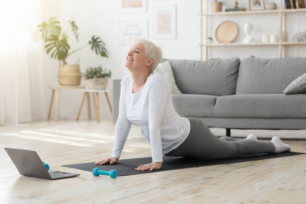 A senior woman doing a cobra yoga pose in her living room.