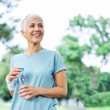 A happy, active older woman smiling outside while holding a water bottle.