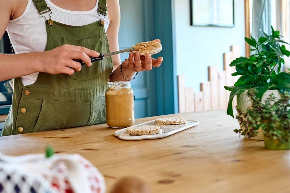 A woman wearing green overalls in the kitchen spreading peanut butter on a muffin.