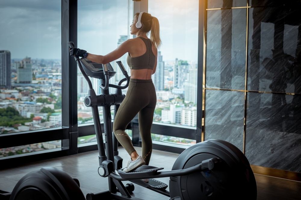 A woman at the gym exercising on an elliptical machine in front of large windows.