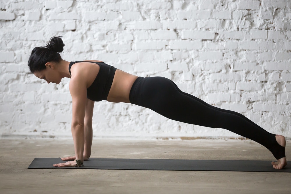 A woman in black workout clothes doing a full-arm plank exercise on a yoga mat.