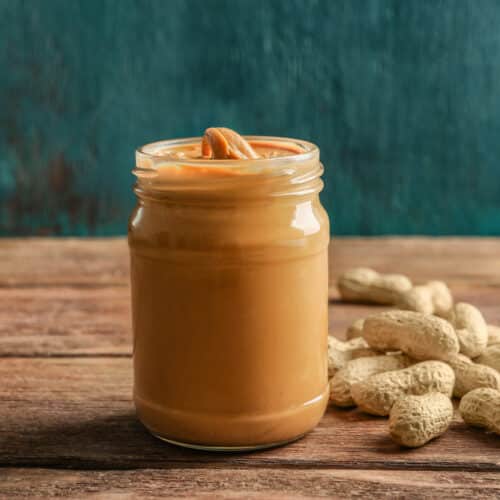 A jar of creamy peanut butter next to peanuts on a wooden table against a painted green background.