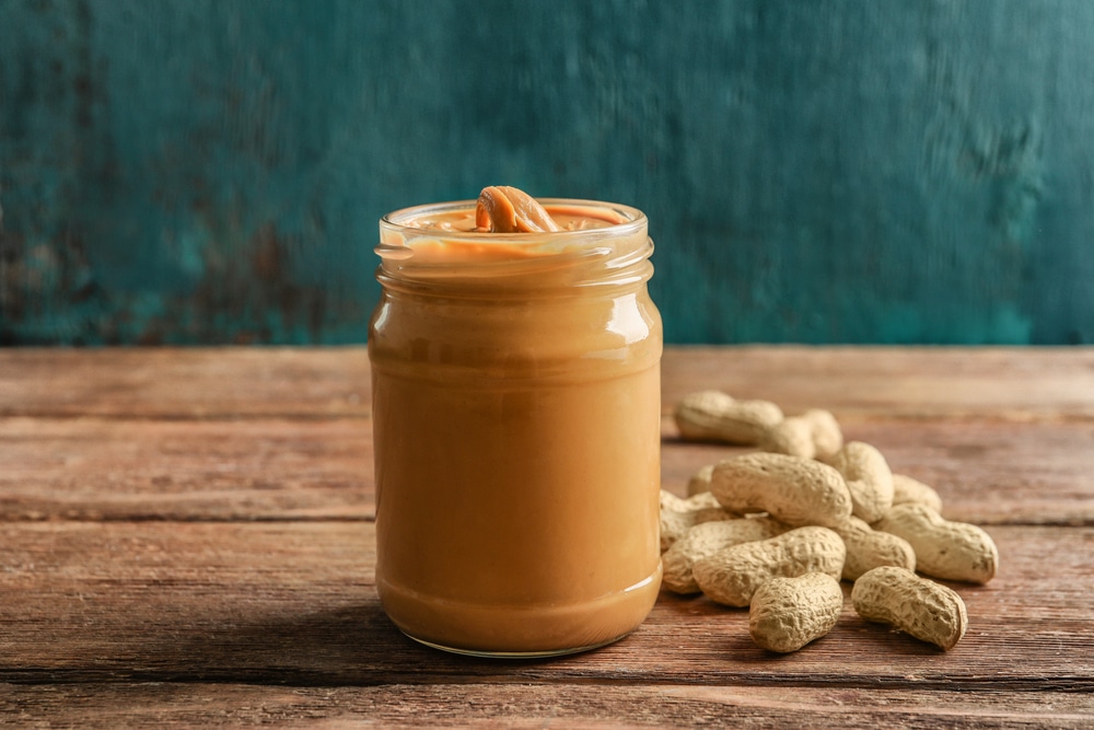 A jar of creamy peanut butter next to peanuts on a wooden table against a painted green background.