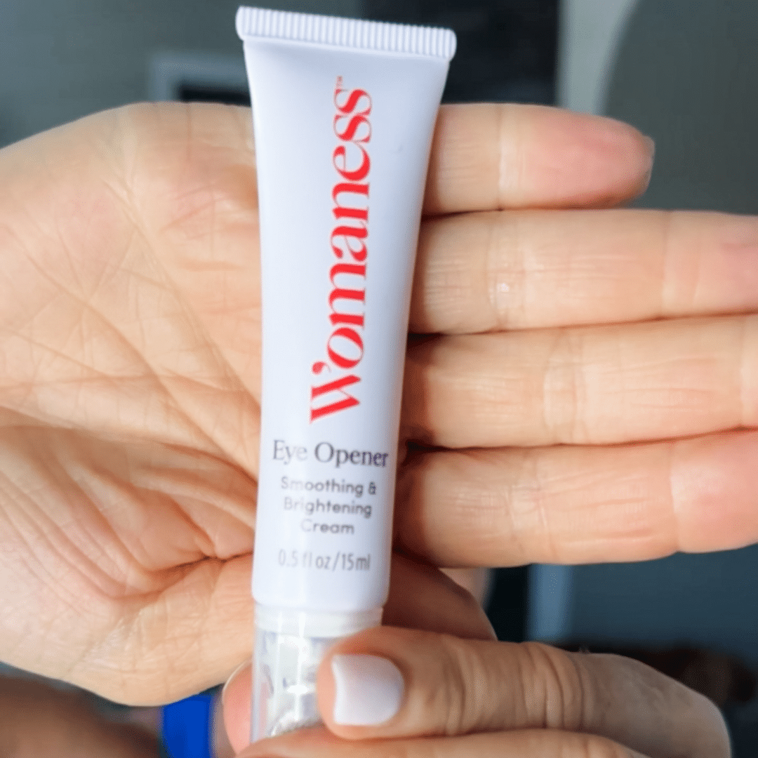 Woman holding up a white bottle of Womaness Eye Opener cream with hand behind the product.