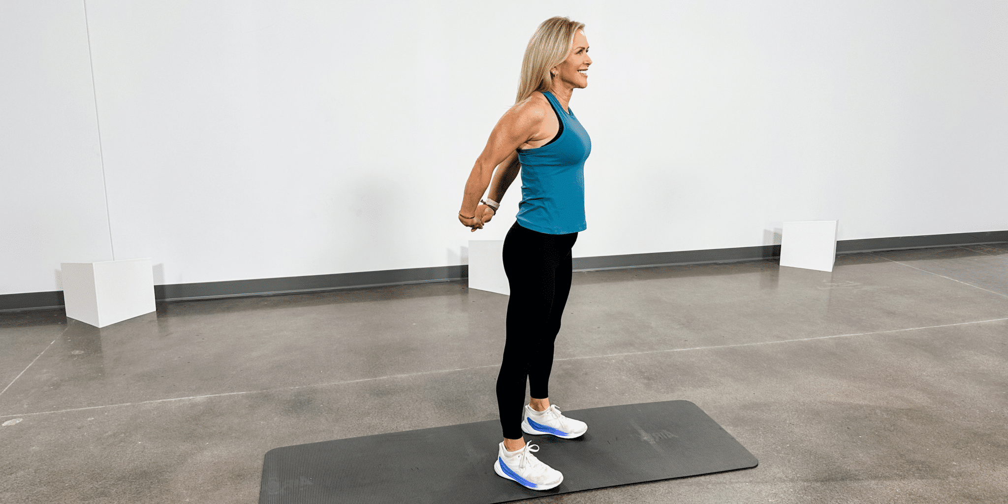 Chris Freytag wearing a blue workout tank and black leggings doing a standing chest expansion.