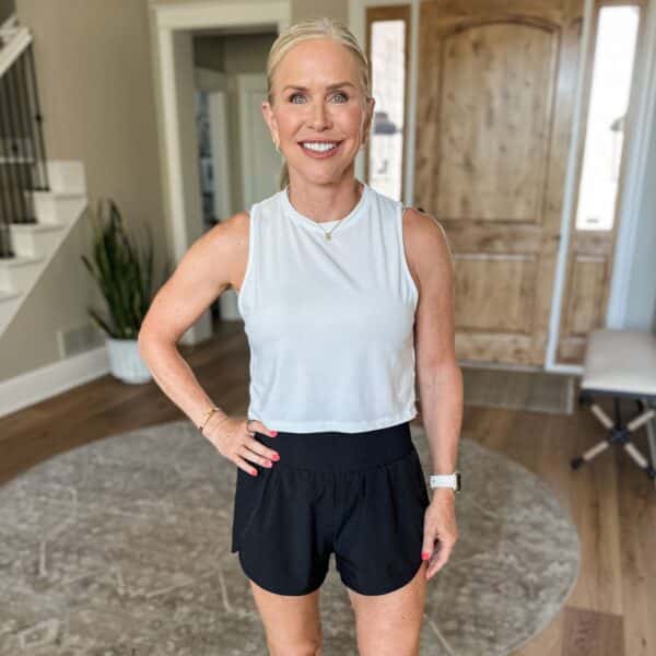 A smiling woman with blonde hair in a ponytail stands indoors on a rug, wearing a white sleeveless top, black shorts, a watch, and jewelry. A wooden door, stairs, and a potted plant are visible in the background.