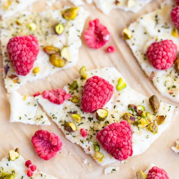 Close-up of pieces of frozen yogurt bark topped with fresh raspberries, chopped pistachios, and herbs, arranged on parchment paper.