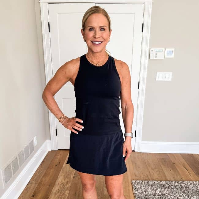 A woman stands indoors on a wooden floor, smiling at the camera. She is wearing a sleeveless black top and a matching skirt, with her left hand on her hip, in front of a white door and beige walls.